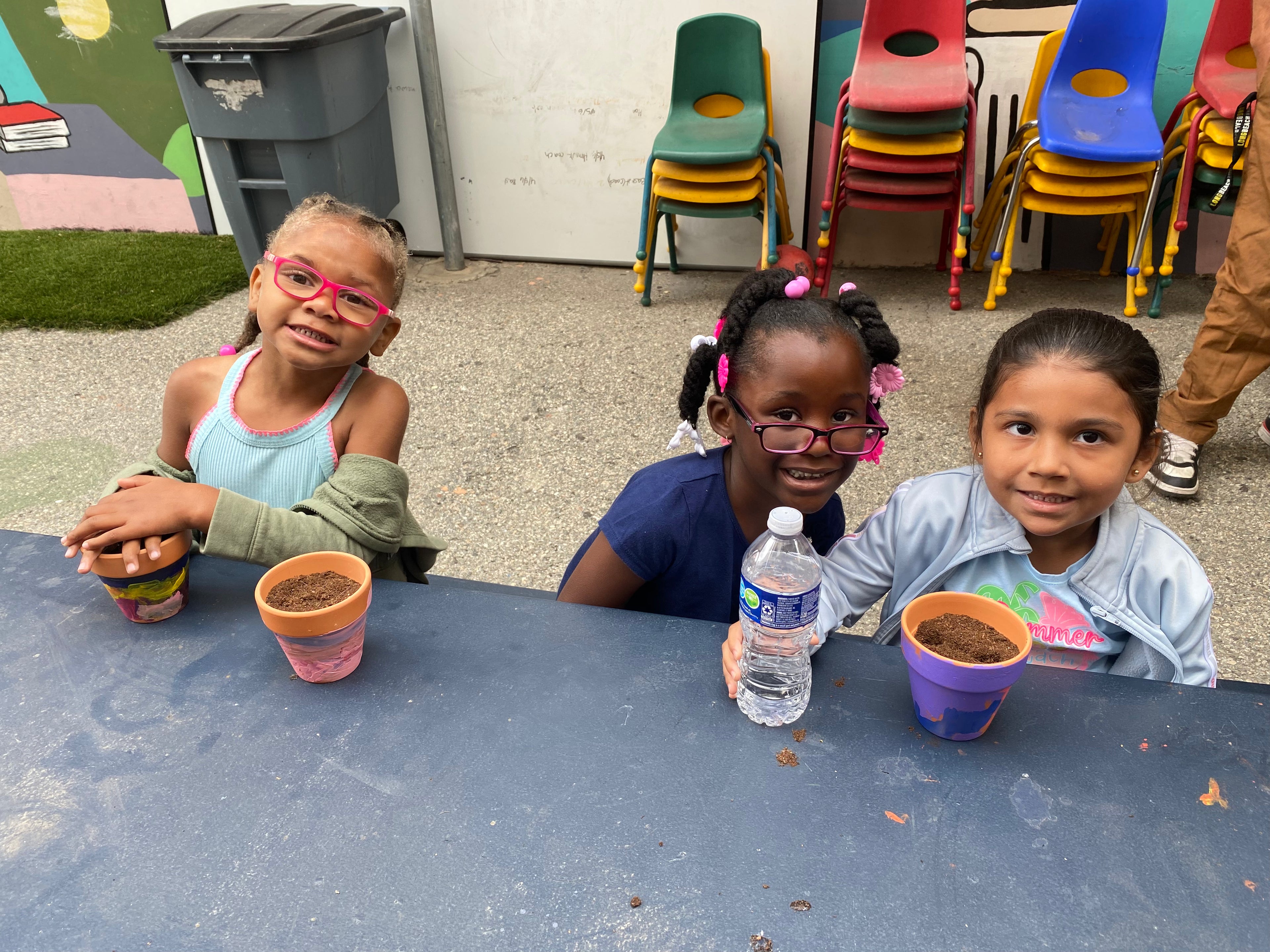 Three children sitting at a table with small pots and a bottle, with colorful chairs in the background.